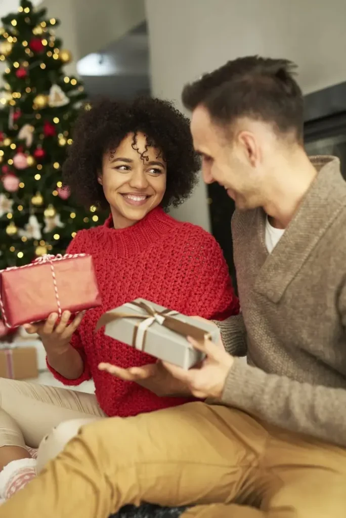 Casal sorridente troca presentes de Natal em frente a uma árvore decorada e iluminada.