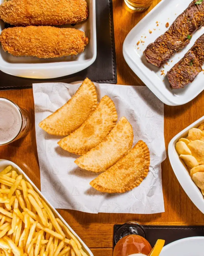 Quatro pastéis fritos dourados sobre papel branco em uma mesa de madeira, cercados por batata frita, carne grelhada e acompanhamentos.