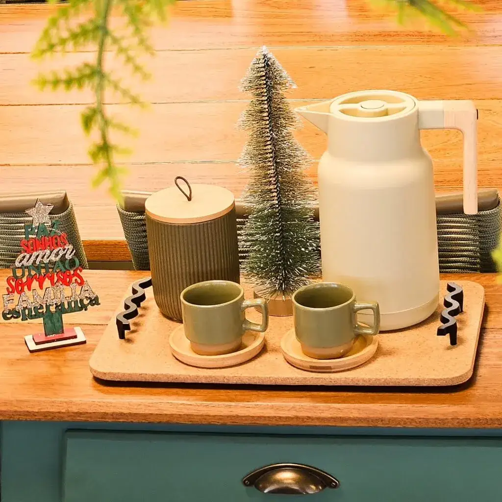 Bandeja com garrafa térmica branca, duas xícaras verdes sobre pires de madeira, pote canelado e pequena árvore de Natal.