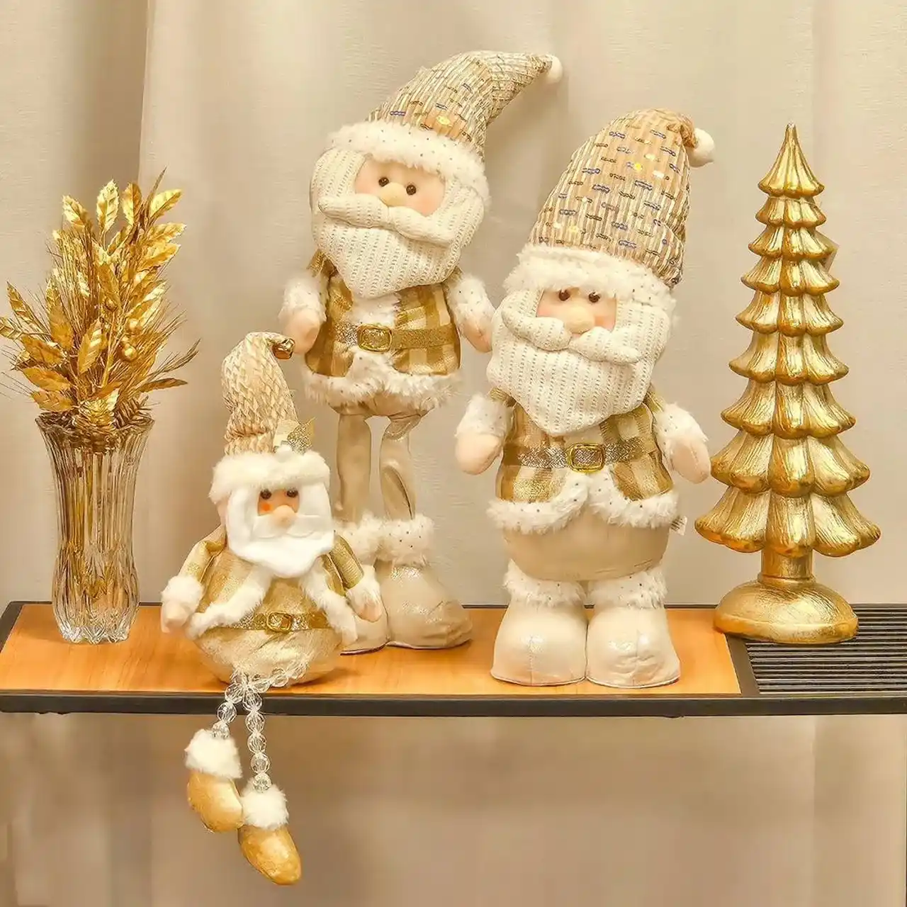Três bonecos de Papai Noel decorativos em tons dourados e creme, ao lado de uma árvore de Natal e um vaso dourados.