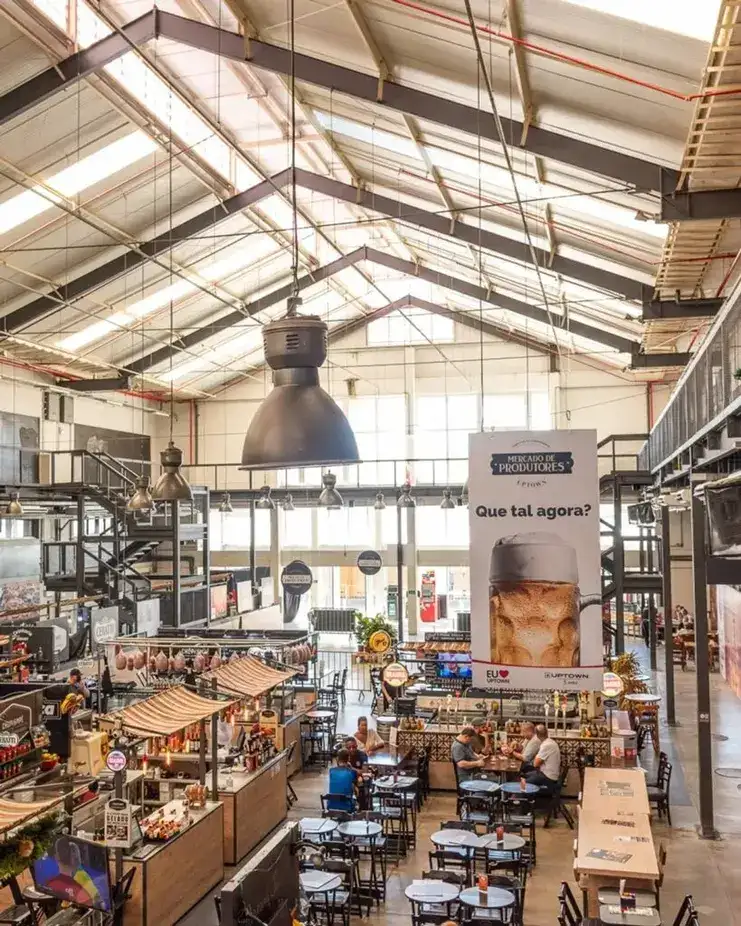 Vista aérea do Mercado de Produtores Uptown com mesas, quiosques de alimentação e teto industrial iluminado por claraboias.