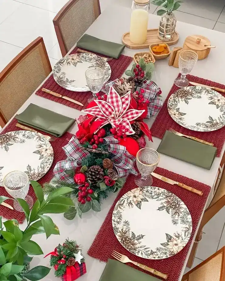 Mesa de Natal com pratos florais, jogos americanos bordô, guardanapos verdes e arranjo central com poinsétia xadrez.