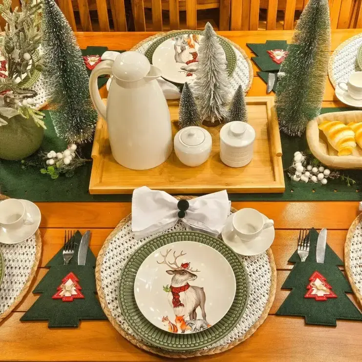 Mesa de Natal com pratos de rena e raposa, sousplats de palha, garrafa térmica branca e mini árvores de pinheiro.