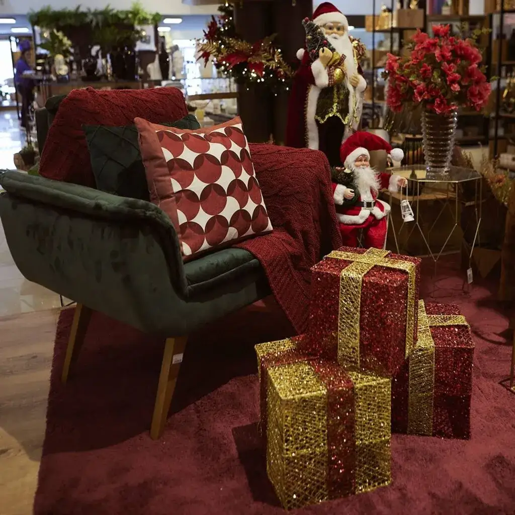 Poltrona verde com manta bordô, almofada geométrica, caixas de presentes brilhantes e Papais Noéis decorativos ao fundo.