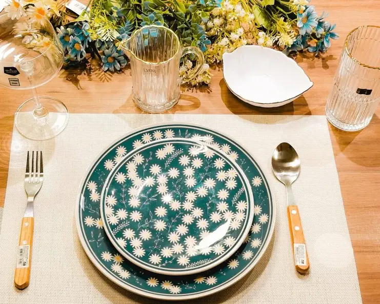 Conjunto de pratos decorados com margaridas em fundo verde escuro, acompanhados de jarra de vidro e flores secas ao fundo.