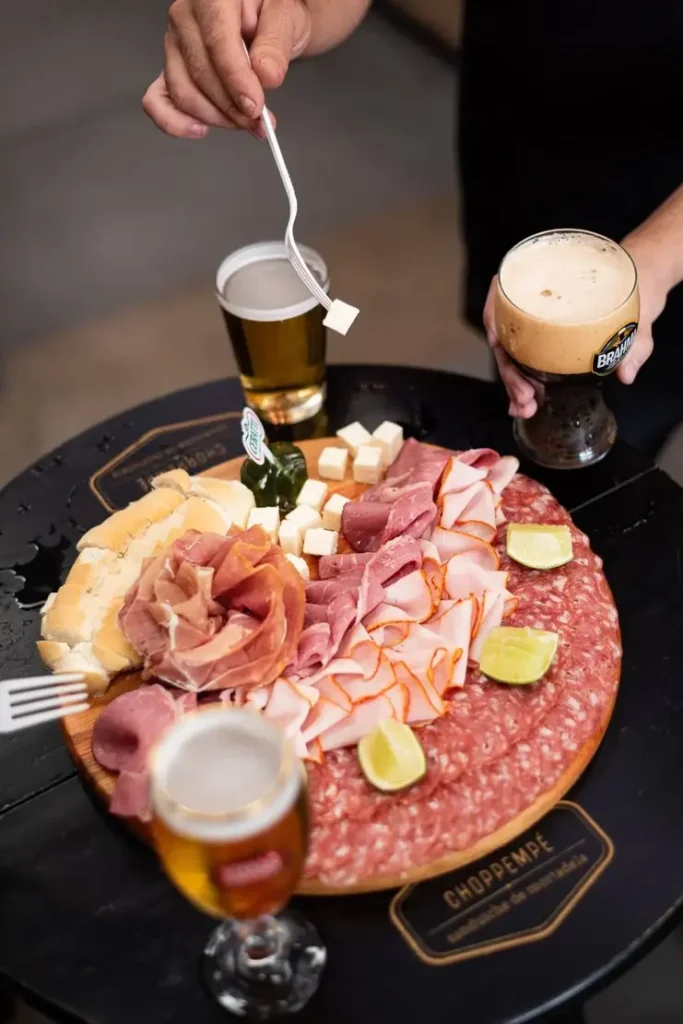 DSC 8275 Choppempe Tábua de frios com salame, presuntos, queijo, pão e limão cercada por copos de chope em mesa de bar.