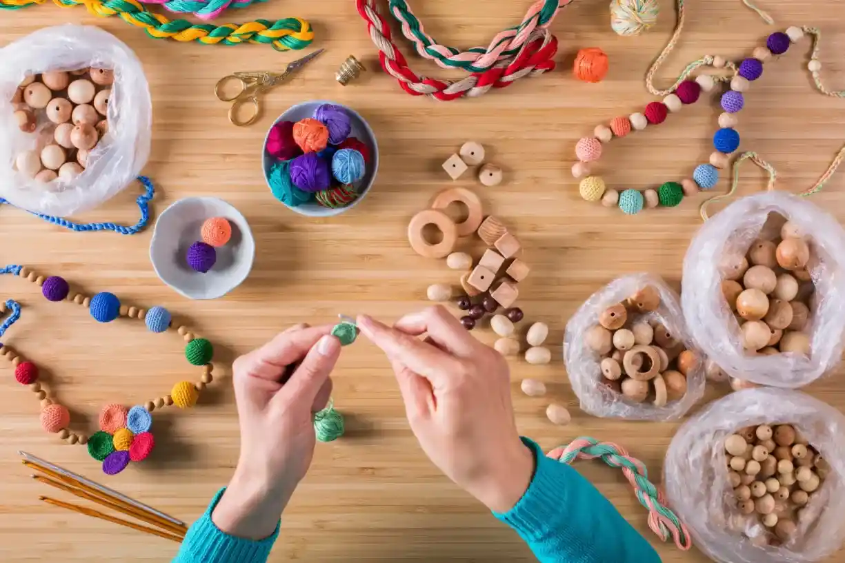 Mãos de artesã fazendo crochê em mesa de madeira repleta de contas, novelos coloridos, agulhas e colares prontos.