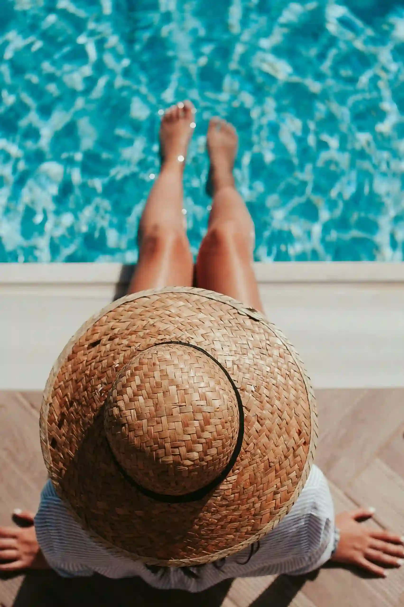 Foto de vista superior de uma mulher com chapéu senta na borda de uma piscina com os pés na água.
