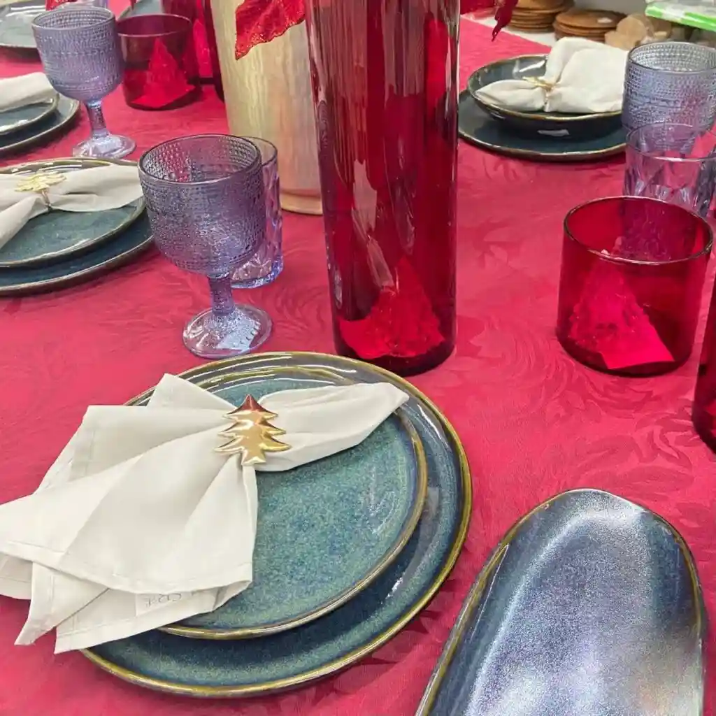 Mesa posta com toalha vermelha trabalhada, pratos em cerâmica verde-escura, guardanapos brancos com anéis dourados em formato de árvore e taças lilases.