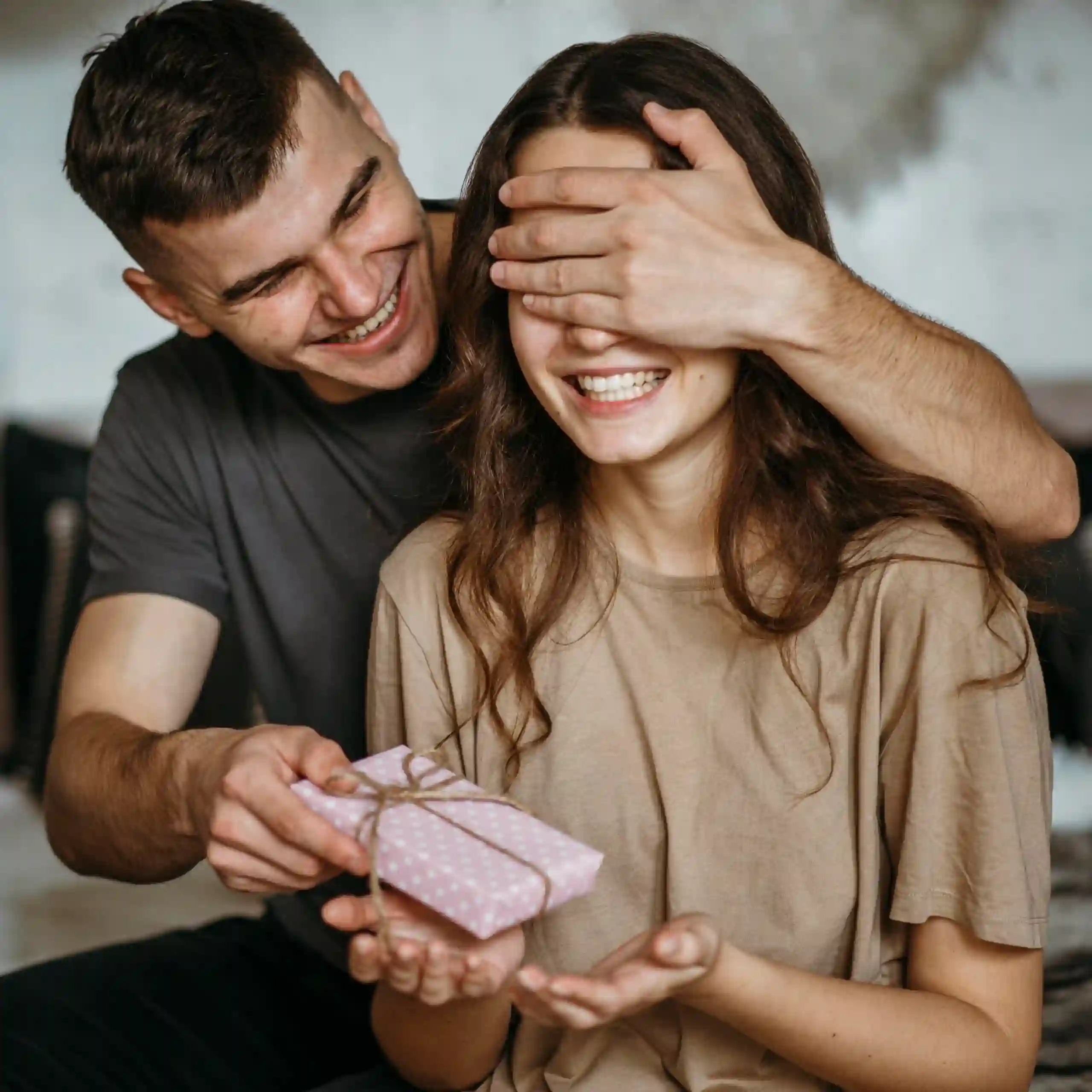 Homem sorridente cobre os olhos de uma mulher e lhe entrega um pequeno presente rosa com bolinhas brancas.