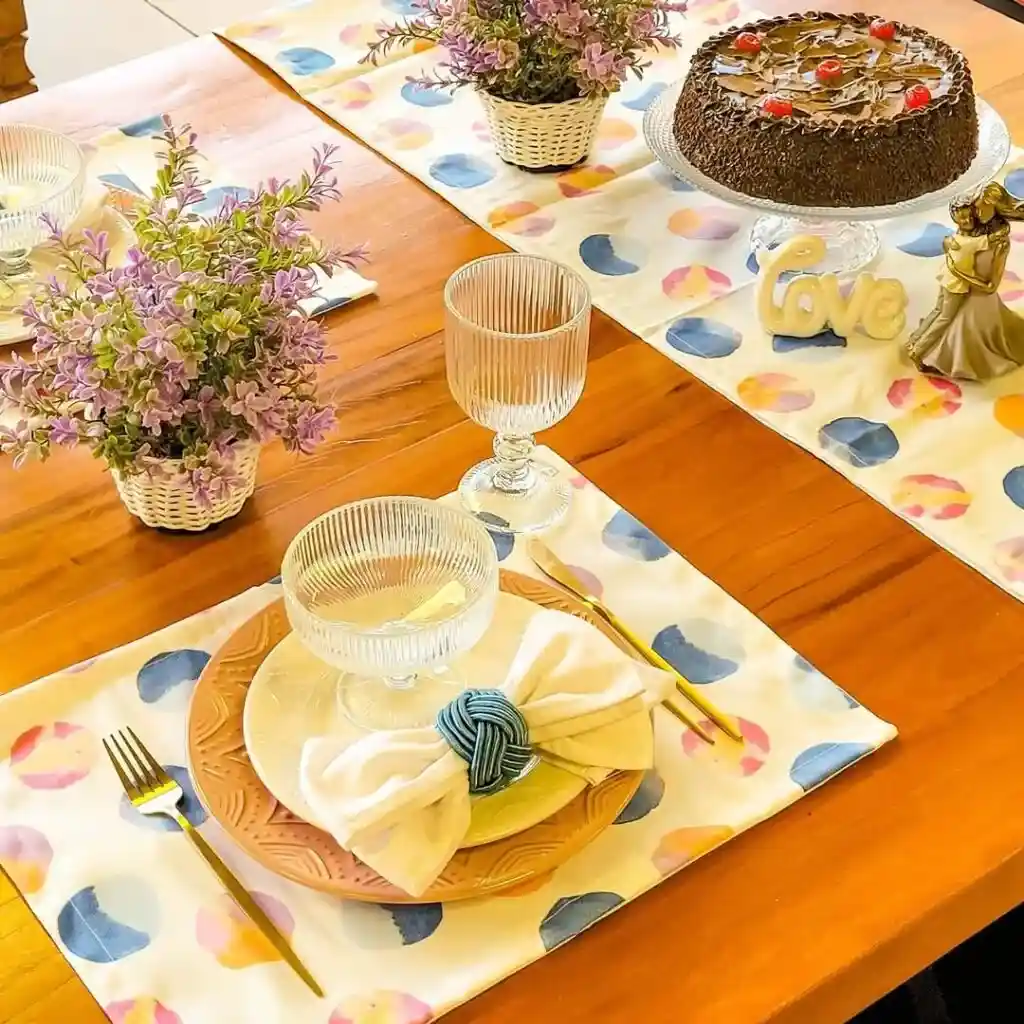 Mesa de madeira com jogo americano de círculos coloridos, louça terracota, taças caneladas, flores e bolo de chocolate.