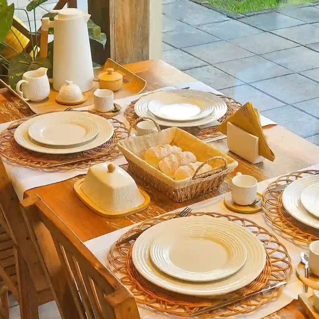 Mesa de madeira posta para o café com louça branca texturizada sobre sousplats de palha trançada. O centro da mesa exibe uma cesta de vime com pães, uma manteigueira branca com base de madeira, um porta-guardanapos com guardanapos amarelos e uma garrafa térmica branca. O ambiente sugere uma varanda, com vista para um piso exterior pavimentado e um pouco de vegetação ao fundo