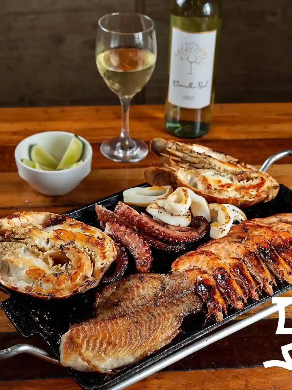 Chapa de frutos do mar grelhados com lagosta, polvo, lulas e camarões acompanhada de vinho branco em mesa de madeira.