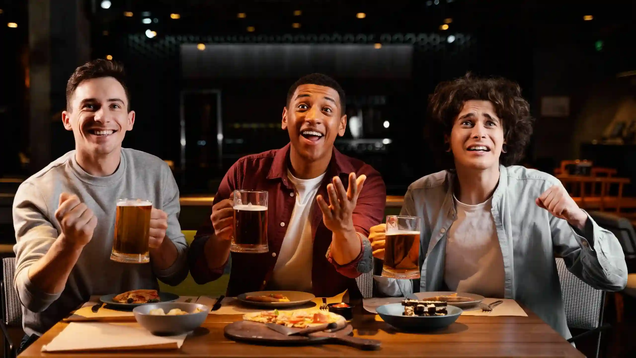 Três homens em um bar comemoram com canecas de cerveja e pizza enquanto assistem TV com expressões de entusiasmo.