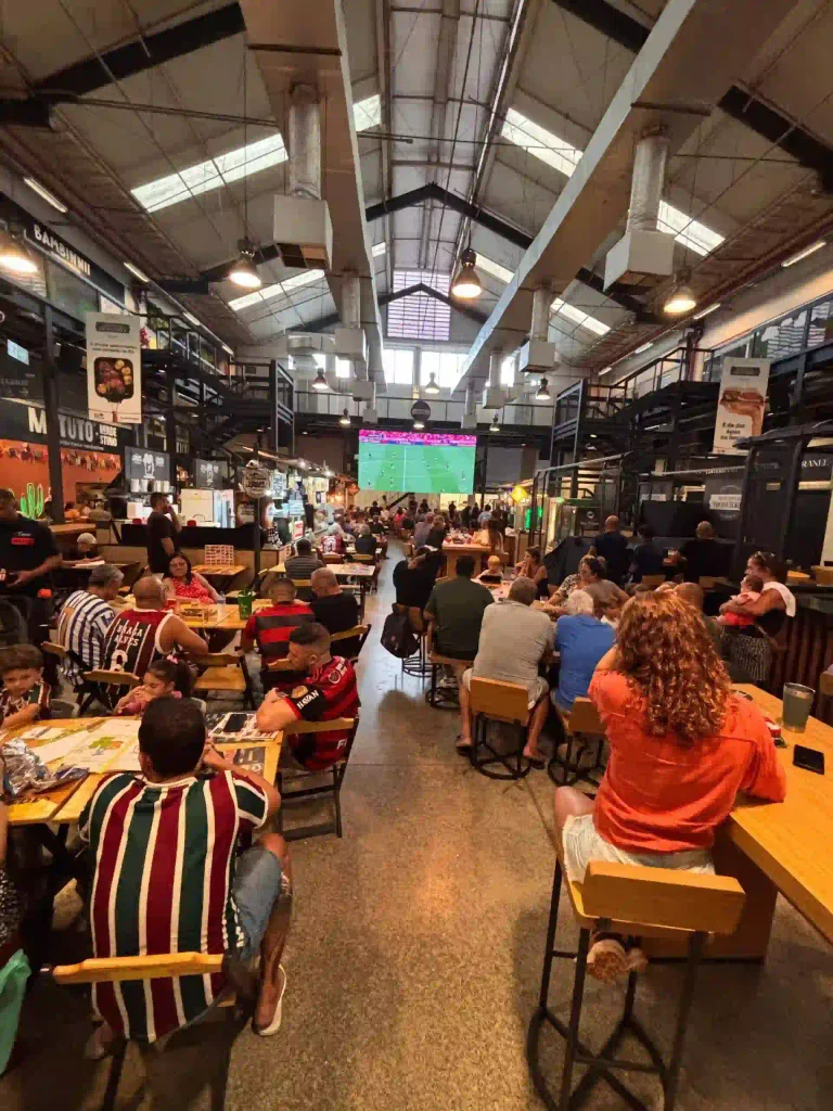 Pessoas sentadas em mesas de um galpão amplo com teto industrial, assistindo a um jogo de futebol em um telão ao fundo.