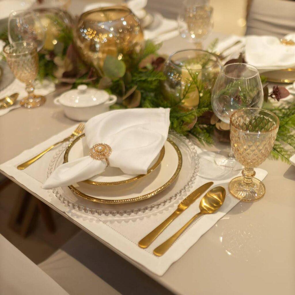 Mesa posta com pratos de cerâmica clara, guardanapos brancos, jogos americanos orgânicos pretos, taças e talheres dourados.