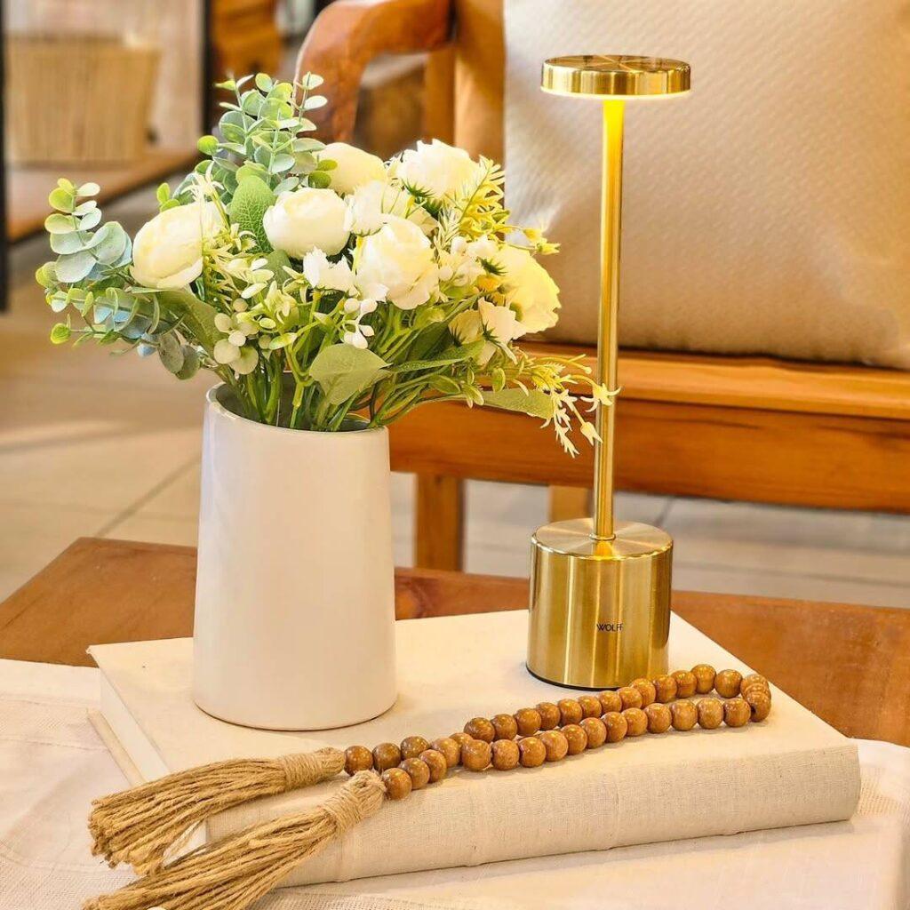 Vaso branco com flores, luminária dourada e terço de madeira sobre livro em mesa de centro.