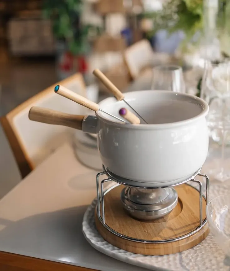 Aparelho de fondue de cerâmica branca com cabo de madeira, sobre base de metal e madeira, acompanhado de garfos com detalhes coloridos.