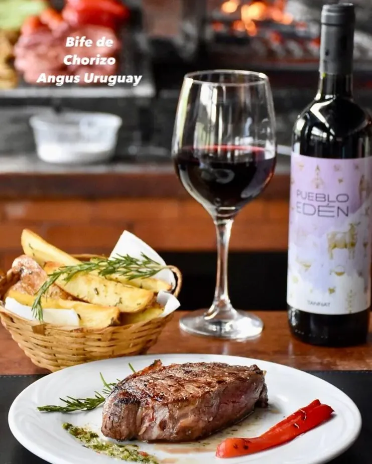 Prato de bife de chorizo Angus Uruguay com batatas rústicas, ao lado de uma garrafa e taça de vinho tinto Pueblo Edén.