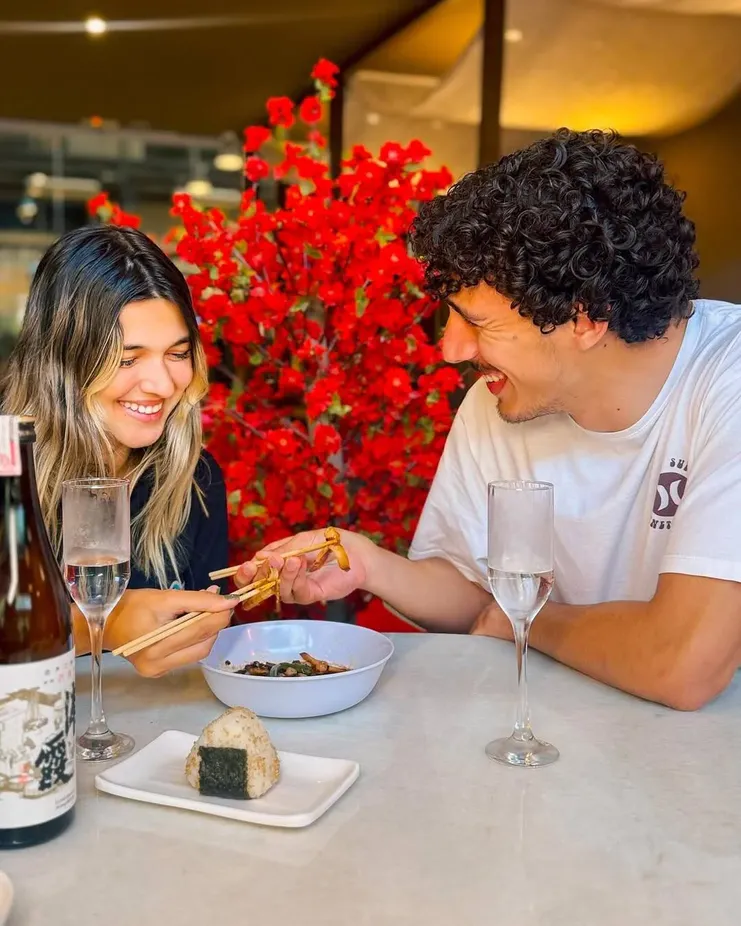 Casal sorridente em restaurante japonês compartilhando comida com hashis; sobre a mesa, onigiri, garrafa de saquê e taças.