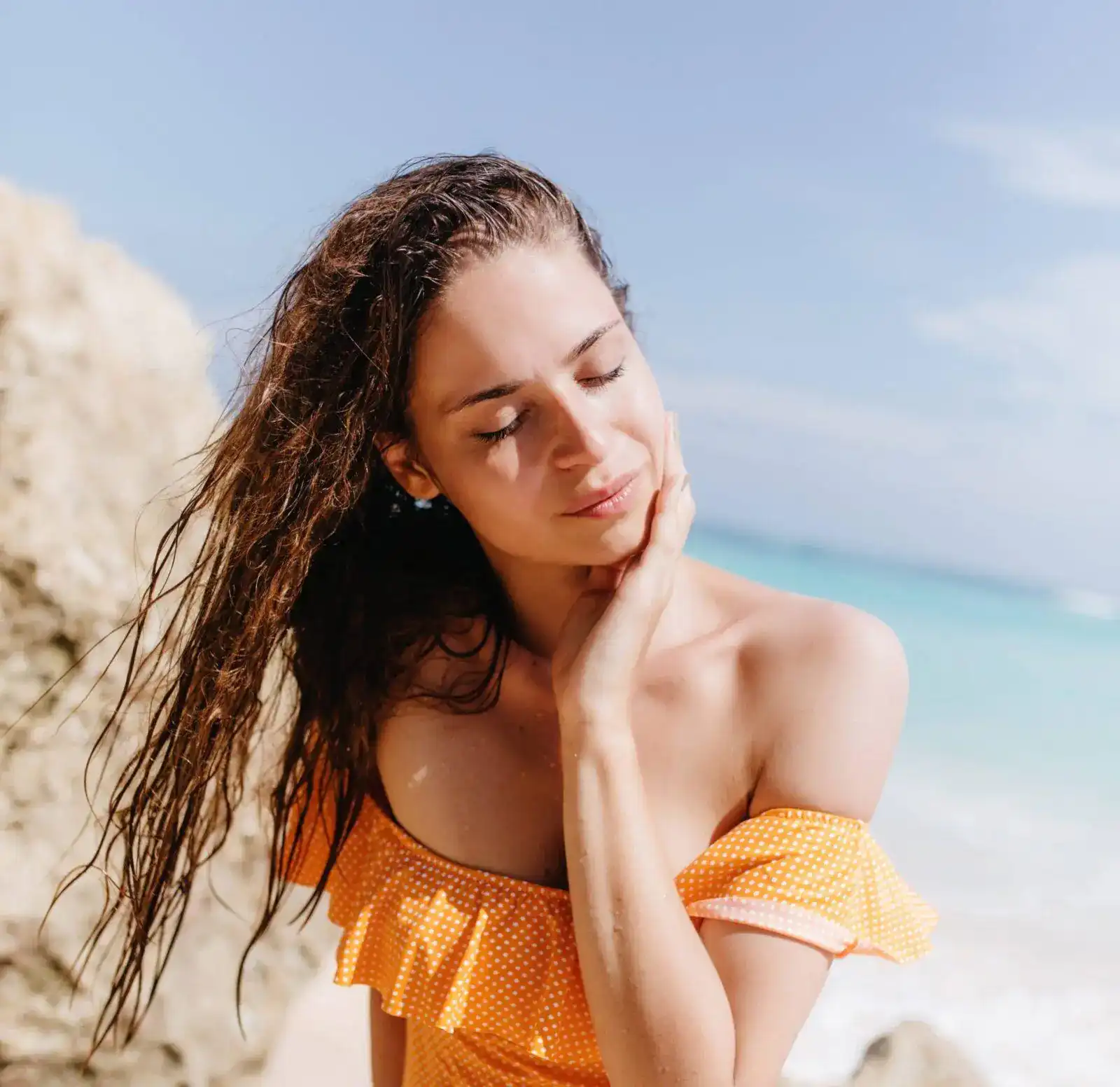 Mulher de olhos fechados com maiô laranja de bolinhas brancas e cabelos úmidos, em frente ao mar sob sol forte.