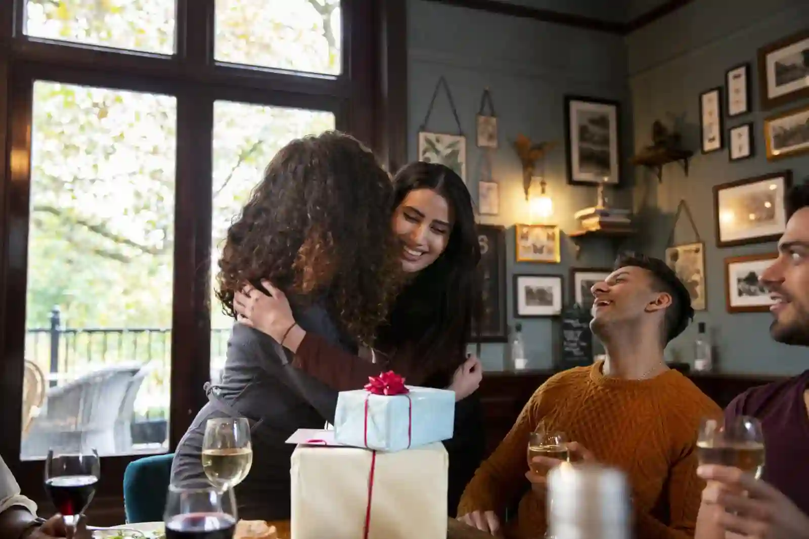Duas mulheres se abraçam em um restaurante, cercadas por amigos que sorriem e brindam com vinho diante de presentes.