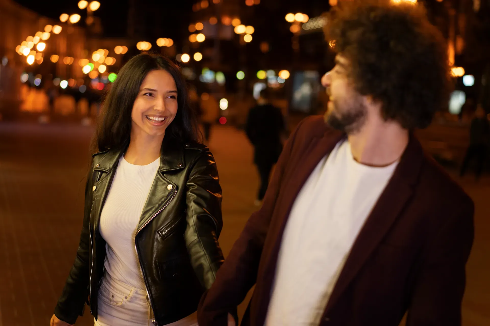 Um casal jovem caminha sorrindo de mãos dadas por uma praça à noite, com luzes da cidade desfocadas ao fundo.