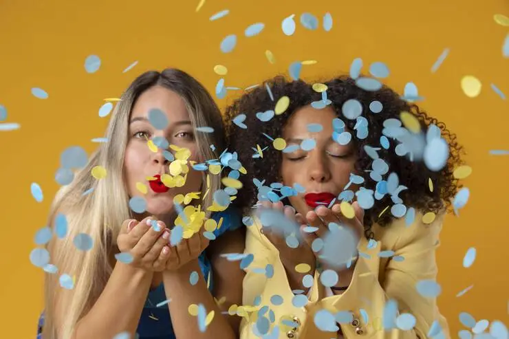 carnavalesquentabailinho-imagebyfreepik Duas mulheres soprando confetes coloridos