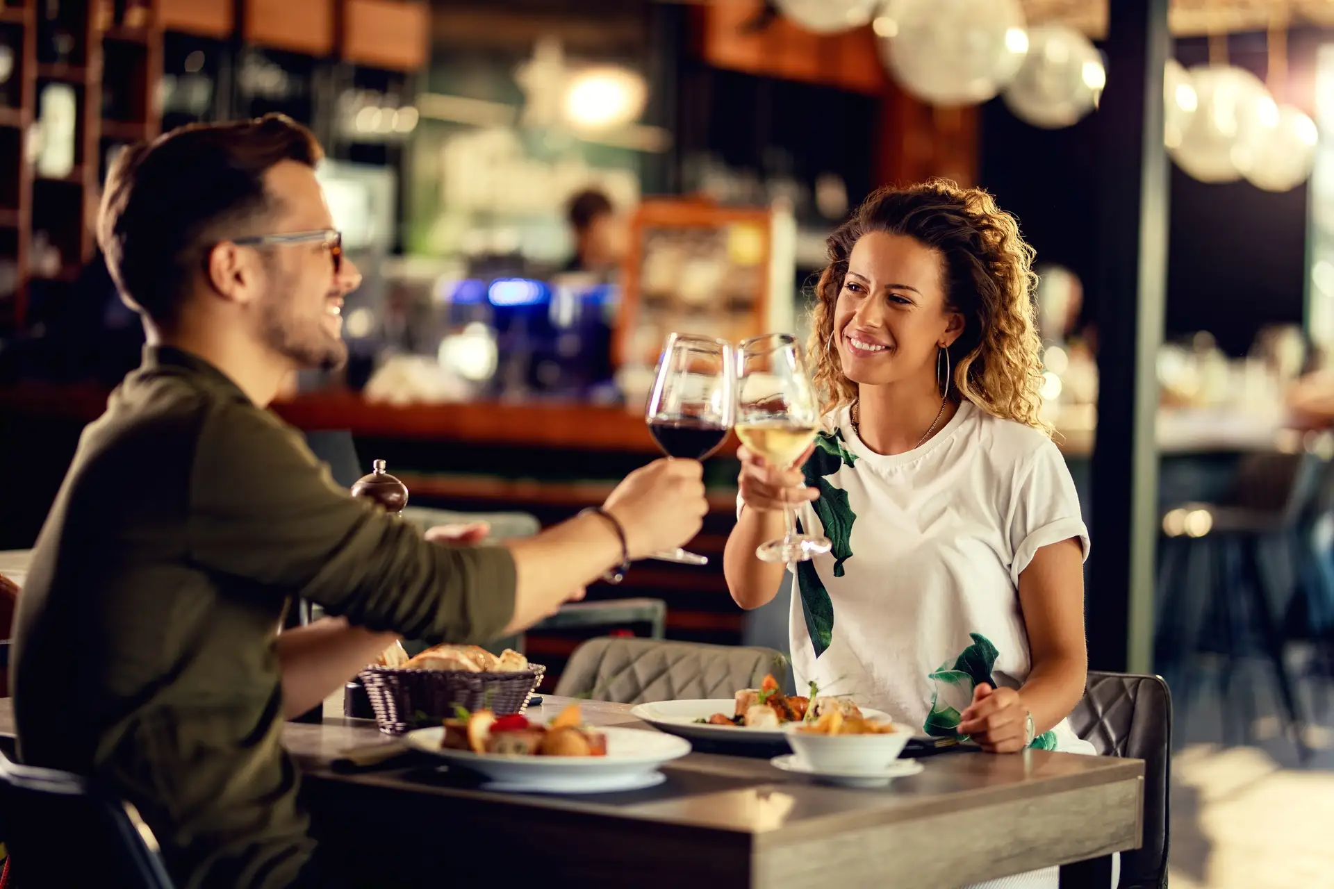 Casal sorridente brinda com taças de vinho durante jantar em restaurante iluminado.