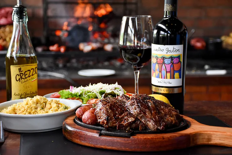 Tábua de carnes e linguiças, arroz, salada, vinho tinto Cuatro Gatos e azeite Il Grezzo com churrasqueira ao fundo.