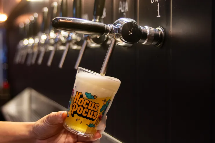 Mão segura copo da cervejaria Hocus Pocus sendo preenchido por chope tirado diretamente de uma torneira de metal.