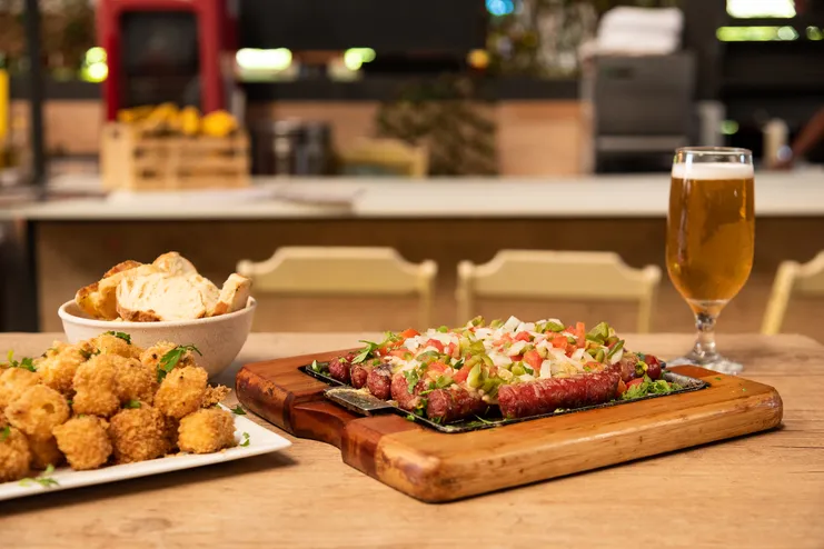 Petiscos de boteco: linguiça acebolada na chapa, dadinhos de tapioca, pão fatiado e um copo de cerveja em uma mesa de madeira.