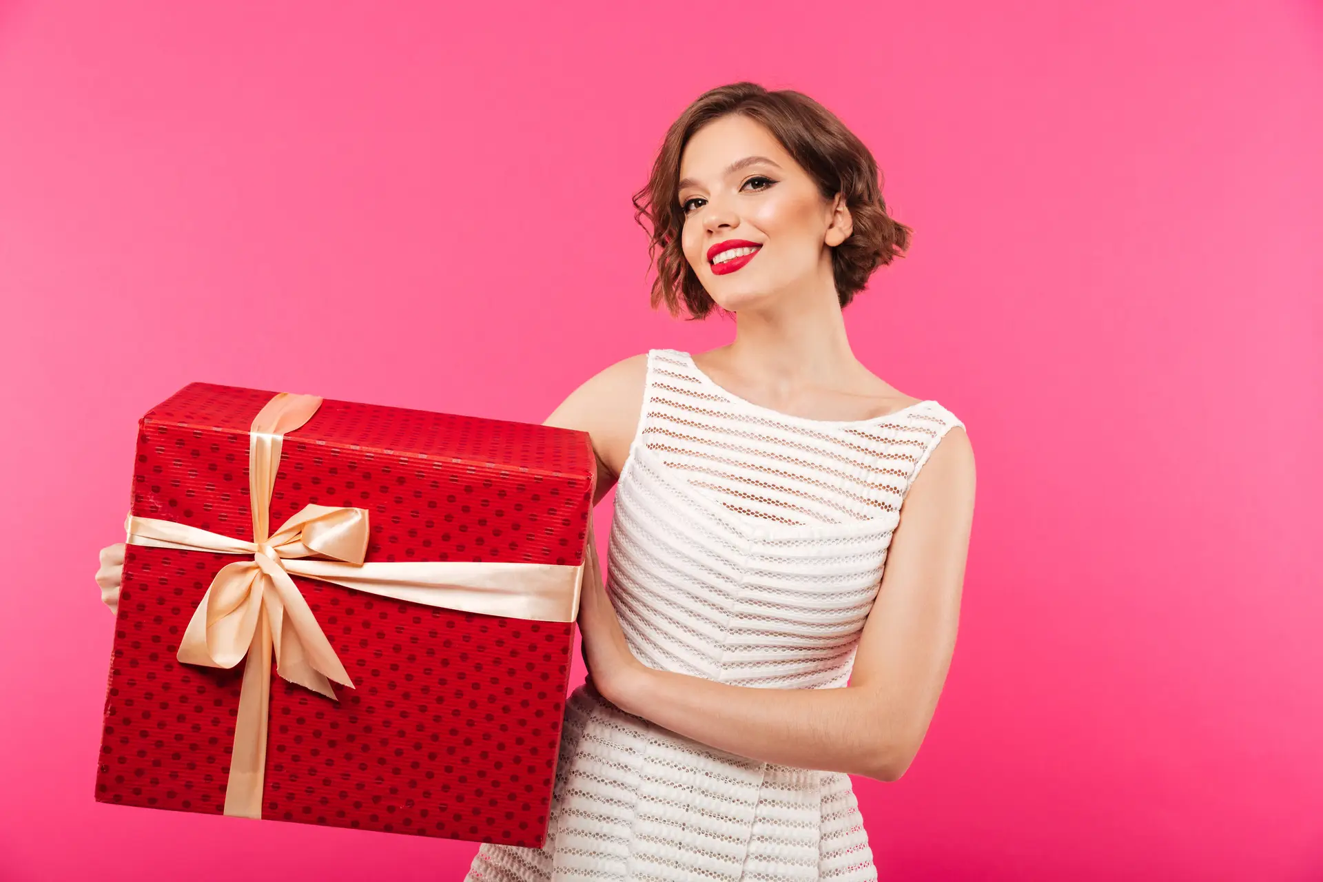 Mulher de batom vermelho e vestido branco segura presente luxuoso, simbolizando festas, aniversários ou promoções especiais.