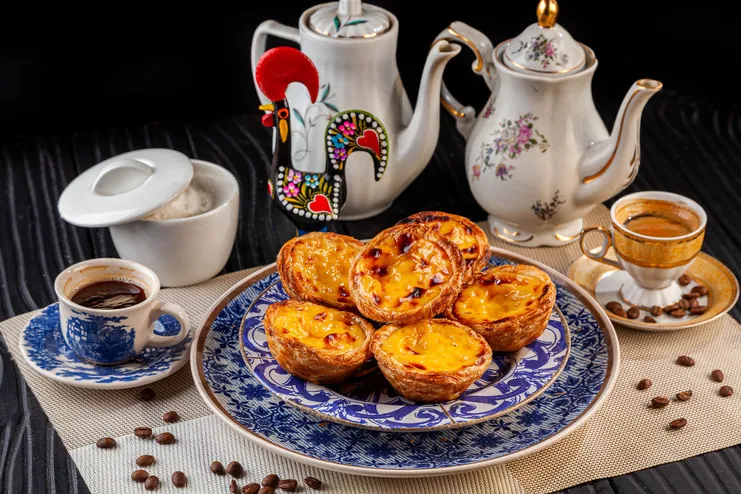 Pastéis de nata em pratos de louça azul, bules de chá, galo de Barcelos e xícaras de café sobre mesa escura.