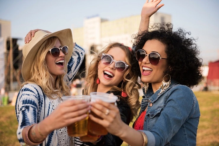 Três amigas sorridentes usam óculos escuros e brindam com copos de cerveja em festival de música ao ar livre.