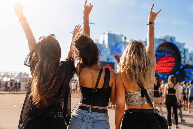 Três amigas de costas com braços erguidos fazendo sinal de rock em frente ao palco de festival de música no sol.