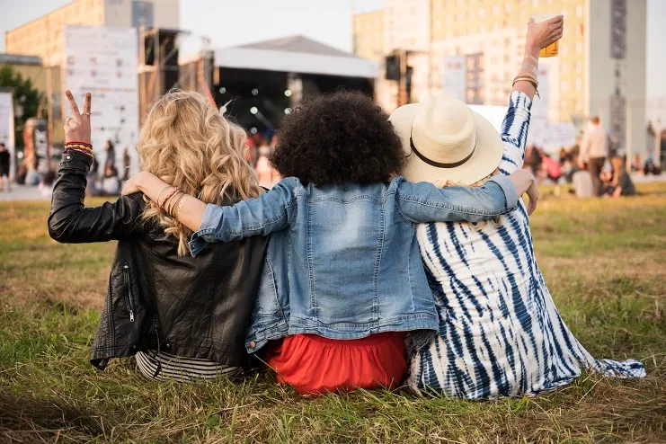 amigos-divertindo-rock-in-rio Três amigas sentadas de costas no gramado de um festival de música, abraçadas, celebrando com um brinde e sinal de paz em frente ao palco.