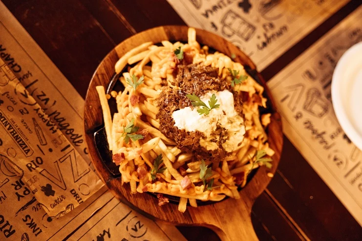 Batata frita com queijo derretido e costela desfiada servida em chapa de ferro com tábua de madeira em mesa de bar.