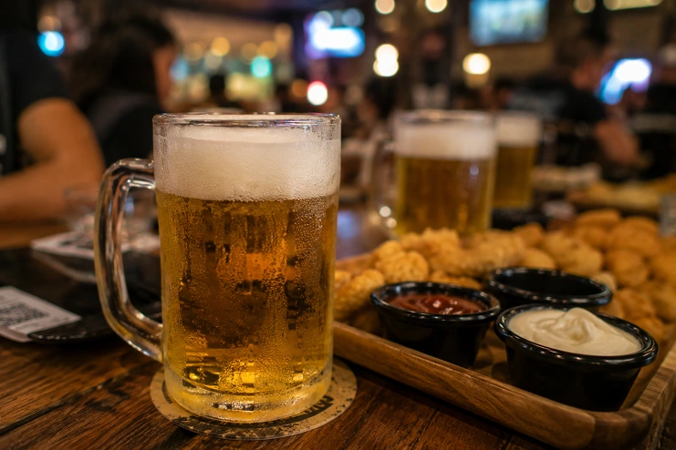 Caneca de chopp gelado com colarinho de espuma ao lado de porção de petiscos fritas com molhos em mesa de bar.