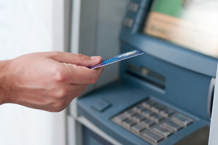 Mão segurando um cartão de crédito azul próximo à entrada de um caixa eletrônico de banco para fazer saque.