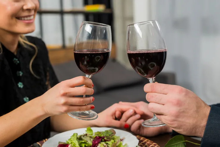 Close de casal brindando com taças de vinho tinto sobre mesa com salada; mãos dadas ao fundo.