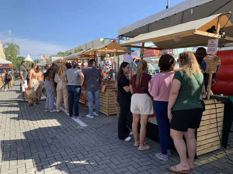 Visitantes em fila diante de barracas de madeira com toldos bege em feira ao ar livre durante o dia.