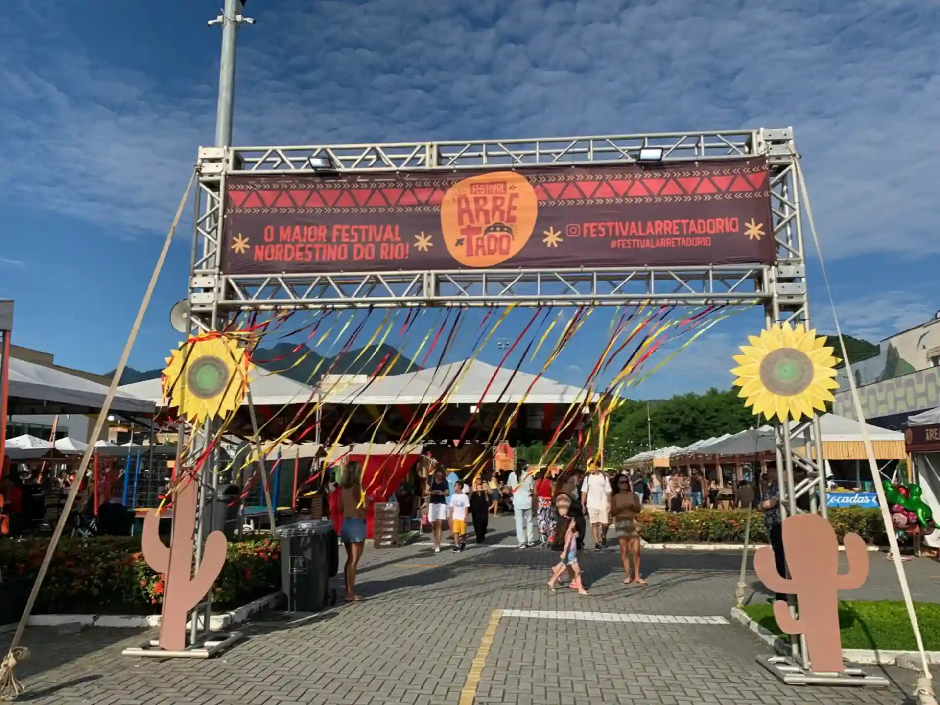 Pórtico de metal com banner do Festival Arretado, decorado com girassóis de papel, cactos e fitas coloridas.