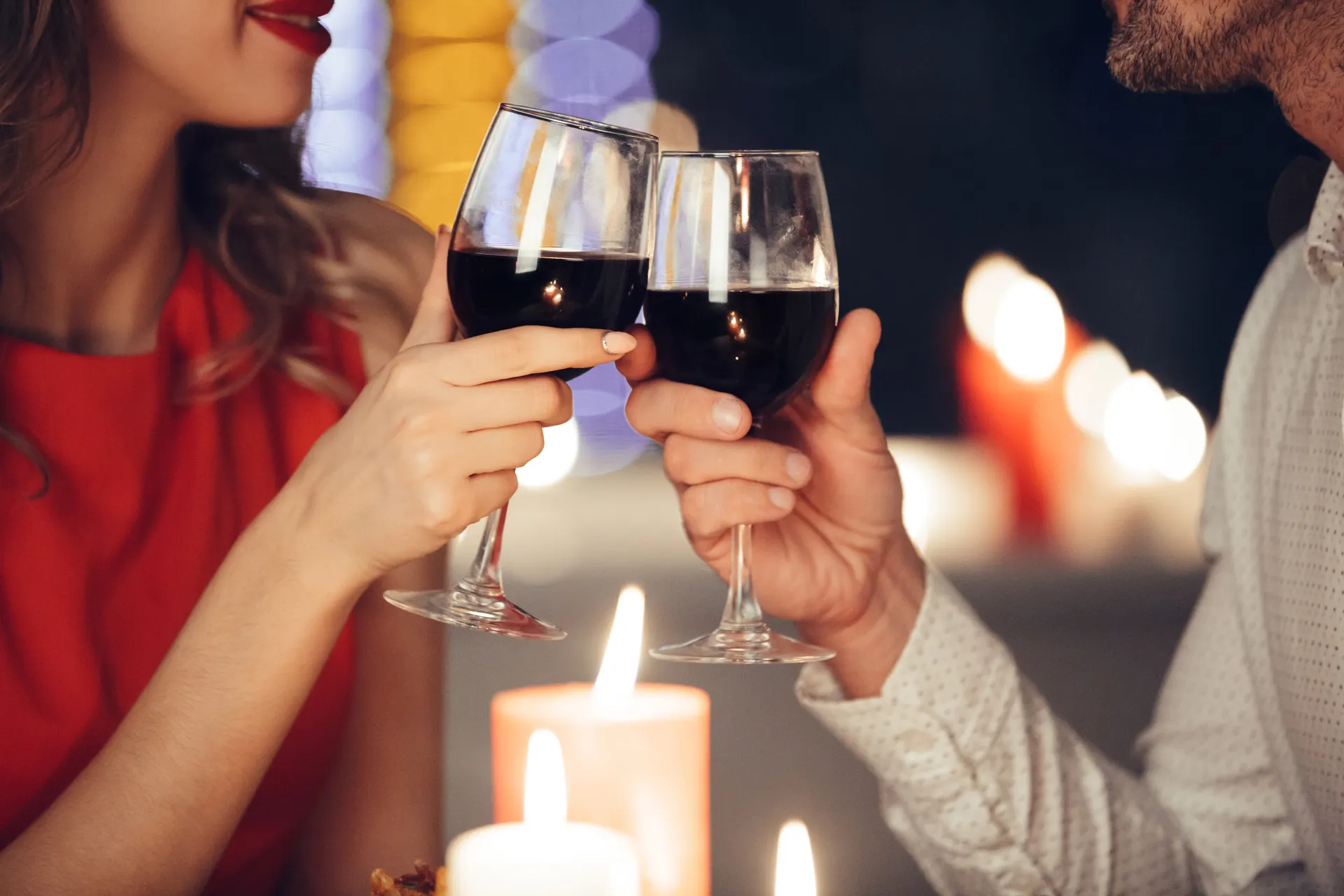 Casal brinda com taças de vinho tinto em mesa iluminada por velas, fundo com luzes bokeh desfocadas.