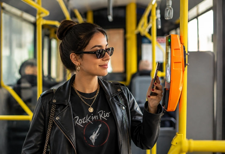 Jovem com jaqueta de couro e camiseta do Rock in Rio aproxima celular para pagar passagem em validador de ônibus.