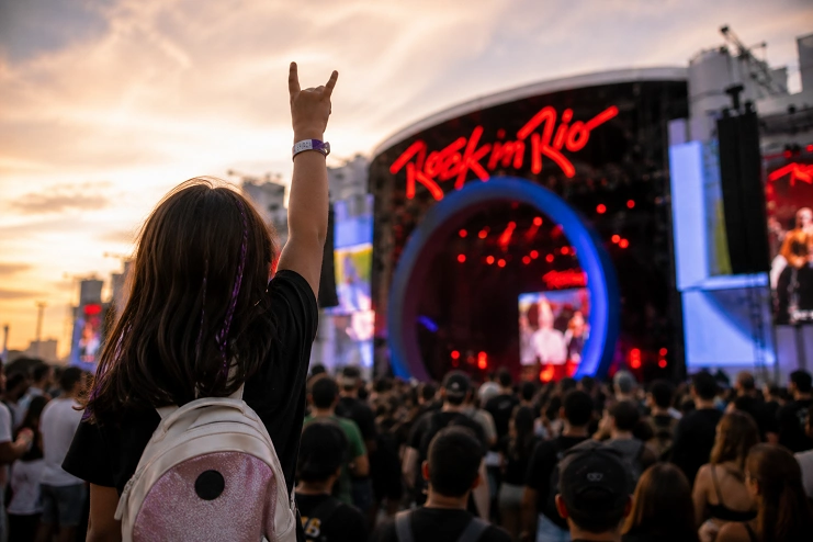 Jovem de costas com mochila faz sinal de rock em frente ao Palco Mundo no festival Rock in Rio durante o pôr do sol.