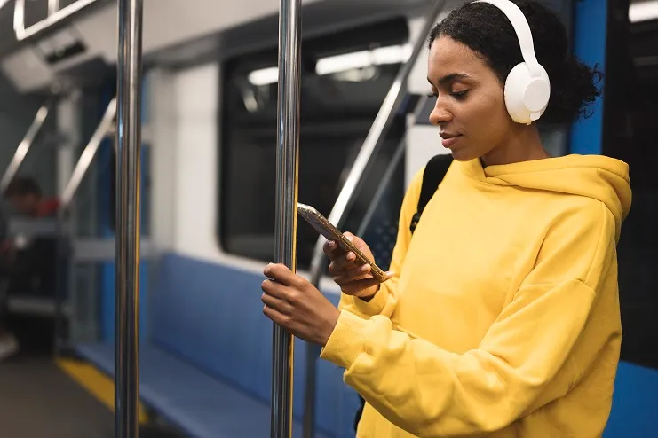 Mulher negra de moletom amarelo e fones brancos usa celular no metrô, segurando-se em uma barra metálica do vagão.