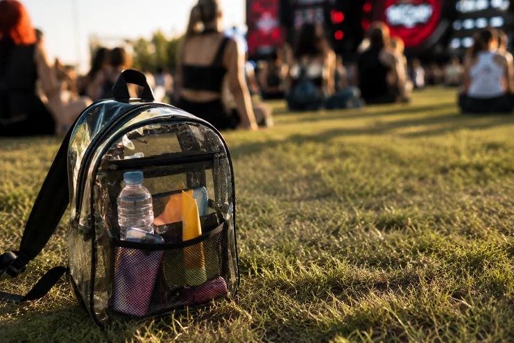 Mochila transparente com garrafa de água e protetor solar no gramado em frente ao palco de festival de música.