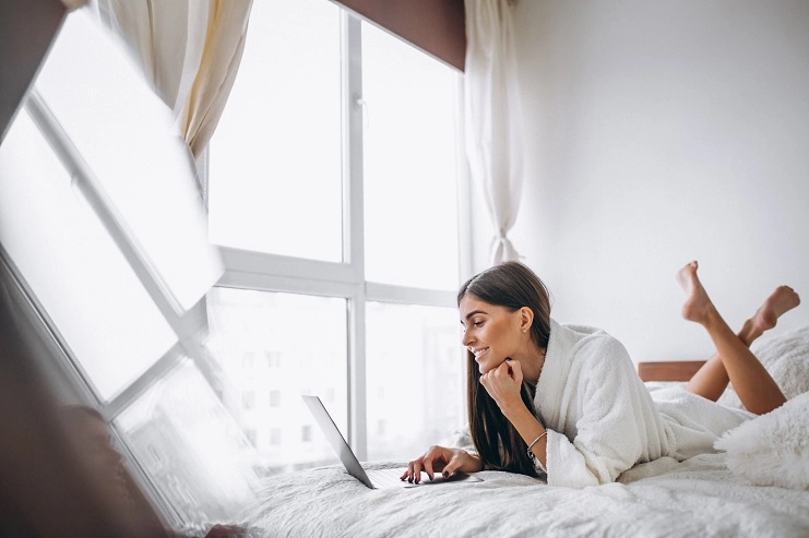 Mulher sorridente de roupão branco deitada na cama usando notebook em quarto iluminado por uma grande janela.