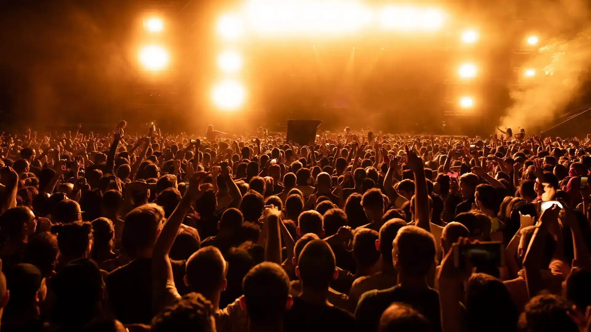 Multidão de costas com braços erguidos diante de palco iluminado por fortes luzes laranjas em festival de música.